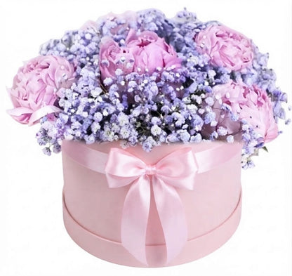 A round pink gift box filled with a bouquet of light pink peonies and lavender baby's breath, tied with a pink satin bow, on a white background.