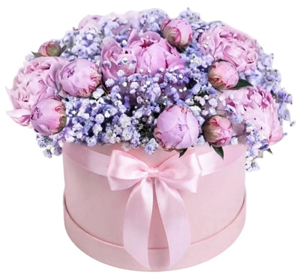 A round pink gift box filled with a bouquet of light pink peonies and lavender baby's breath, tied with a pink satin bow, on a white background.